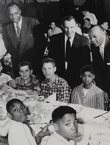Olympic gold medalist Jesse Owens, top right, leans over a young Junior Watkins at a banquet honoring the Peoria Little League, February 1957. Standing next to Owens are Henry Harper, Carver Center executive director, and coach Tony Van Dyke of Roosevelt Junior High School.
