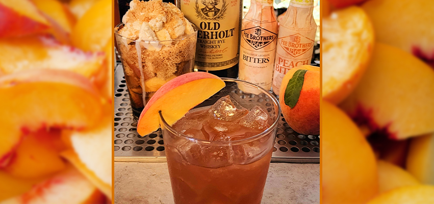 A cocktail glass with a peach wedge in front of various bottles of alcohol