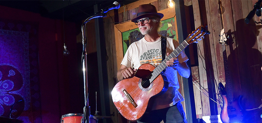 Man holding a guitar on stage with drums in background.